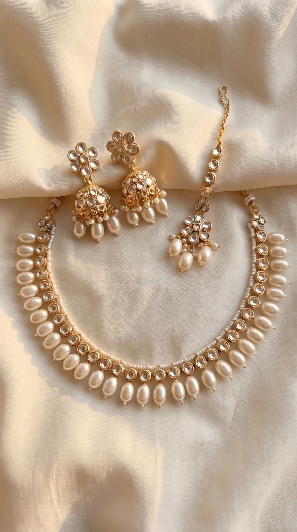 Gold-finish kundan necklace with pearl-drop fringe, matching kundan jhumka earrings with pearl drops, and floral kundan maang tikka, displayed on ivory satin fabric.