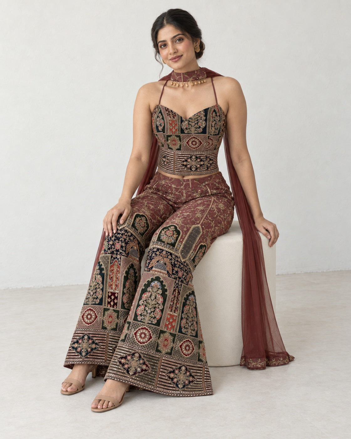 AI-styled model view: Woman seated on an ivory stool, wearing the embroidered top, wide palazzo trousers and scarf.